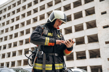 Tablet in hands. Professional firefighter is standing outdoors against big unfinished building