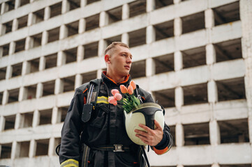 Beautiful flowers in hands. Professional firefighter is standing outdoors against big unfinished...