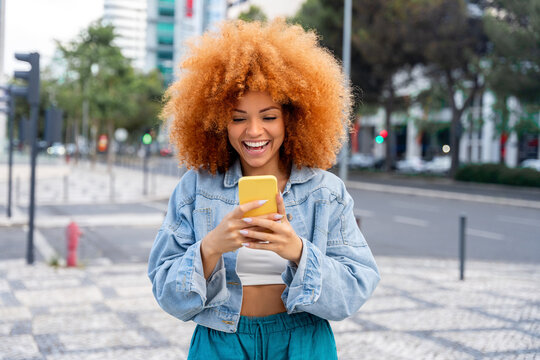 Happy excited young African american woman shopper winner holding cell phone outdoors at city street. Latin female student girl feeling joy using mobile app, winning gift, bonus, reading news online