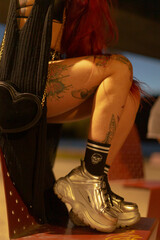 A tattooed woman with vibrant red hair sits in a stylish chair, wearing fashionable silver sneakers...