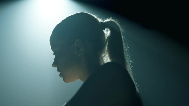 Side view of a woman cycling in a dark studio, backlit with soft cinematic light as the camera moves to the front, revealing her focused eyes and deep breathing during intense training effort