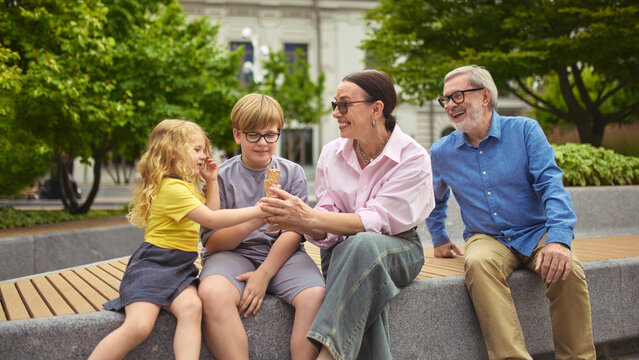 Grandparents and grandchildren sitting on bench with ice cream outdoors. Concept of summer leisure, family bonding, park treat, outdoor eating, lifestyle generation bridge.