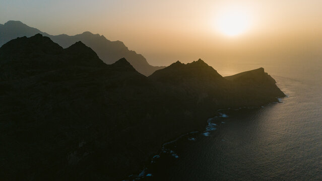 Aerial view of rugged, shadowed peaks meet the shimmering ocean under a hazy sun, creating a striking contrast of light and dark, Mirador del Balc&Atilde;&sup3;n, Canary Islands, Spain.