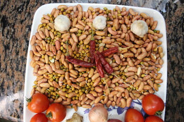 Raw, uncooked ingredients for a healthy meal displayed on a white platter. Includes sprouted beans, fresh vegetables like tomatoes, onions, ginger, garlic, and dried red chilies.