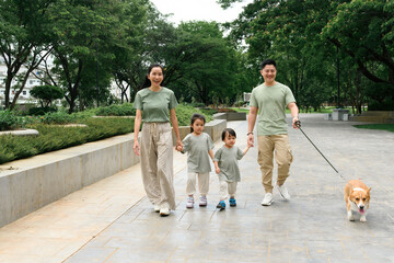 An Asian Family of four enjoys a relaxing walk through a city park with their cheerful Corgi,...