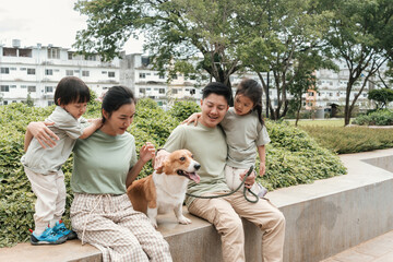A cheerful family of four sitting on a concrete bench with their Corgi, sharing time in a peaceful...
