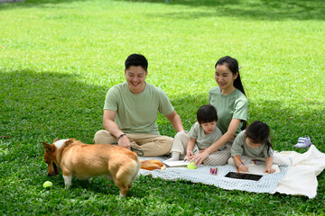 Smiling family enjoying a casual picnic on the grass with their children and cheerful corgi dog