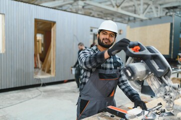 Indian carpenter cuts a wooden plank on a sawmill in a modular building factory, providing precise...