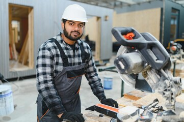 Indian carpenter cuts a wooden plank on a sawmill in a modular building factory, providing precise...