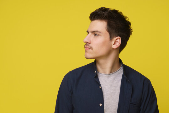 A young man with dark hair and a mustache looks thoughtfully to the side, standing against a bright yellow backdrop. He is wearing a navy blue shirt over a gray t-shirt - Powered by Adobe