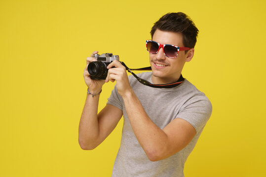 A man wearing sunglasses holds a camera up to his eye and smiles, while standing in front of a solid yellow background
