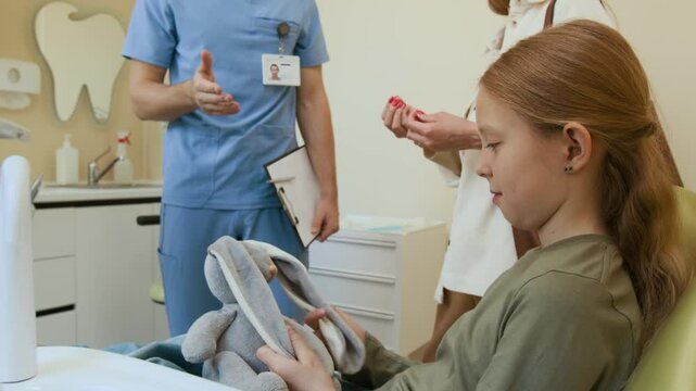 Side view shot of smiling little girl playing with toy bunny while sitting in dental chair, unrecognizable dentist talking to parent in background