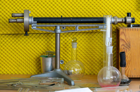 old analytical balance surrounded by glass laboratory vessels and test tubes, placed on a wooden desk against a yellow wall background &mdash; scene from a vintage chemistry laboratory.