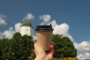 Brown paper cup of drink in women's hand on against backdrop of island Olaf's Tower in town Vyborg...