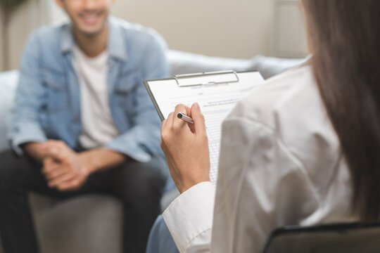 Psychology, depression. Happy smile asian young man patient mental therapy explain symptom with psychologist while doctor woman taking notes at clinic. Psychologist with sick mental health person.