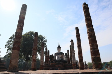Ancient Buddha statue and stone columns at Wat Mahathat, Sukhothai Historical Park, Thailand. Serene heritage site with lush trees and clear sky.