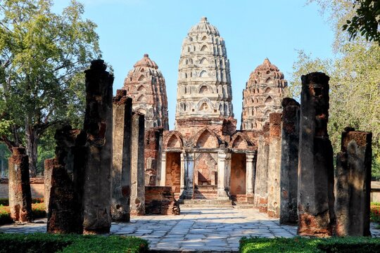 Ancient ruins of Wat Si Sawai in Sukhothai, Thailand. Central prang surrounded by weathered stone columns and lush greenery under a clear blue sky.