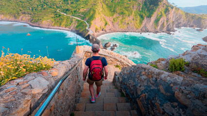 A male hiker with a red backpack descends the famous stone staircase leading to the iconic San Juan de Gaztelugatxe hermitage, a stunning travel destination on the coast of Biscay.