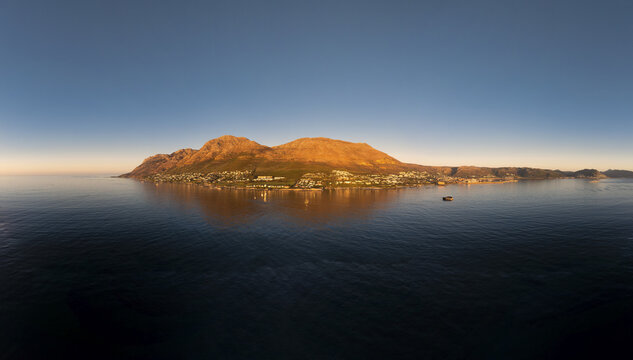 Aerial view of golden light kissing the rugged peaks and tranquil waters surrounding Hout Bay, creating a stunning contrast of warmth and coolness, Cape Town, Western Cape, South Africa.