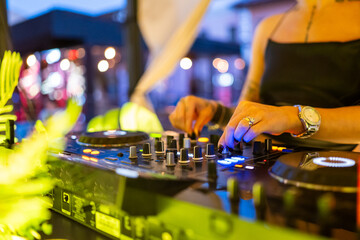 DJ performing outdoors, adjusting mixer controls at vibrant event with colorful lights and energetic atmosphere, focus on hands and professional gear.