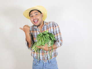 Fototapeta premium Asian male farmer wearing a rattan hat holding a harvested vegetable with a funny expression while pointing to the side