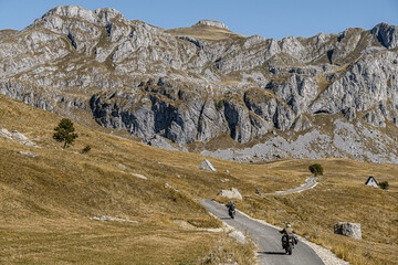 Two motorcyclists ride through a winding mountain road in a rocky, dry alpine landscape under clear skies. Adventure travel, freedom, and scenic exploration theme. Ideal for tourism or nature ads.