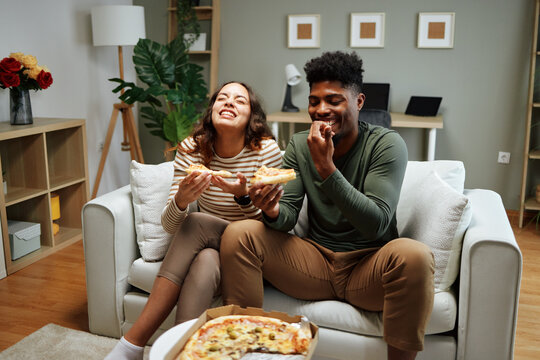 Happy couple laughing and eating pizza on sofa at home - Powered by Adobe