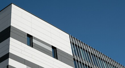 Low angle view of a modern building with geometric shapes, featuring gray and white facade panels under a clear blue sky.