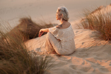 A serene scene of an older woman sitting on sandy dunes, surrounded by tall grasses, absorbing the...