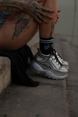 A tattooed leg rests on a stone staircase, showcasing trendy silver sneakers and sleek black socks,...