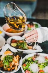 Close-up of a hand holding a glass of white wine over a table with seafood and salads, capturing elegant dining and celebration atmosphere.