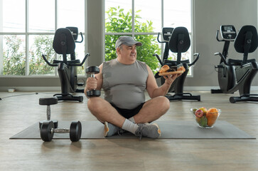 A man sit cross-legged on a gym mat, holding a dumbbell in one hand and a plate of pastries in the other. Exercise bike fill the background, creating a contrast between health and indulgence.