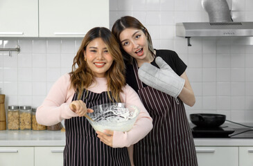 Two women in apron smiling in the kitchen, one holding an oven mitt, the other mixing batter in a...
