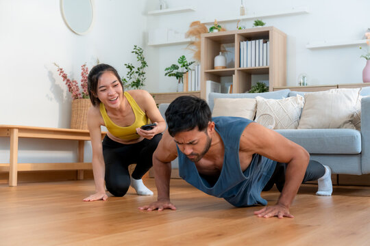 Young asian couple workout during woman hold smartphone encouragement to partner while man doing pushup with motivation, woman cheering man with exercise with strong and strength, self-care. - Powered by Adobe