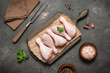 Raw chicken drumsticks on wooden cutting board with spices, meat fork and linen napkin flat lay on dark grunge background, top view