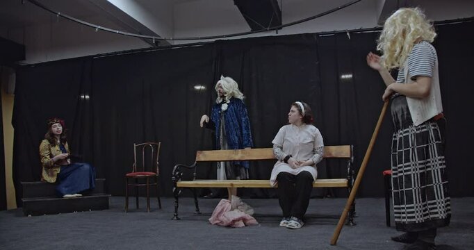 Actors rehearse a play on a stage. A man in a queen's costume and crown gestures while two women look on.