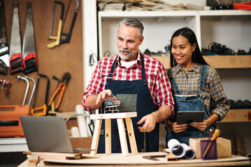 Senior Carpenter and Asian Woman Using Laptop to Sell Handmade Woodwork Products on E-commerce...