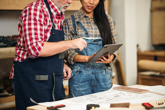 Team of Carpenters Planning Wood Project, Senior Male Craftsman and Young Asian Designer Sketching Furniture Design in Timber Workshop