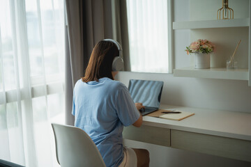 Young woman working on a laptop at a bright home office desk with headphones, surrounded by natural light and modern decor elements, cozy atmosphere