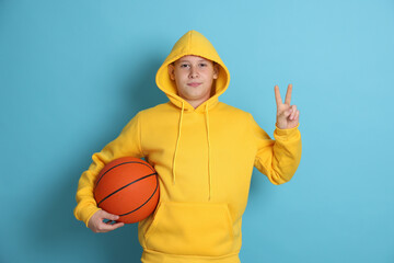 Teenage boy with basketball ball showing peace sign on light blue background