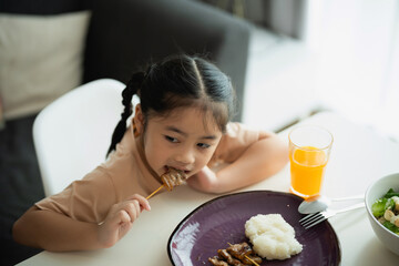 Young child enjoying a meal with rice and meat on a plate, seated at a table with a glass of juice, captured in a cozy dining environment