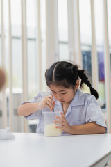 Young girl in school uniform conducting science experiment with glass jar while mixing liquid with spoon in bright classroom environment
