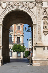 Obraz premium A young, handsome man with a beard, wearing a striped shirt and cargo trousers, rests one leg on a Plateresque and Baroque arch at Seville City Hall in Andalusia. The man is on holiday in the city.
