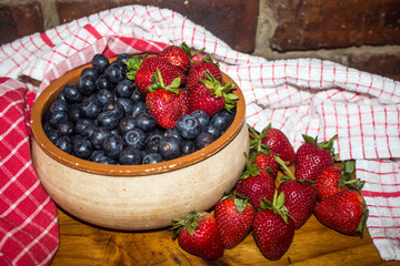 A rustic pottery bowl, filled with Blueberries, surrounded by strawberries and red kitchen Towels