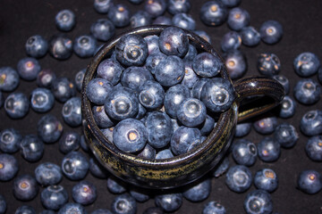 A black ceramic cup, filled to over spilling with large indigo-colored blueberries, with more blue berries surrounding it