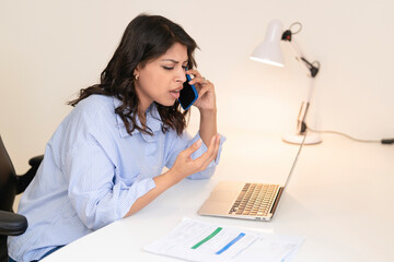 Woman having a serious phone conversation while sitting at a desk with a laptop and documents...