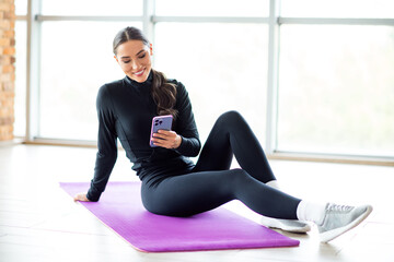 Athletic woman sitting on yoga mat, checking phone during workout in sunny gym, showcasing active...