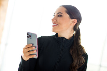 Active young woman holding a smartphone while looking away, dressed in sporty attire in a brightly...