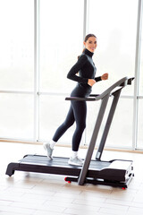 Athletic young woman exercising on a treadmill indoors in a modern gym surrounded by daylight and...