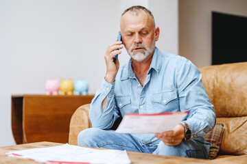 Senior man engaged in a phone call while reviewing important documents at home during a relaxed...